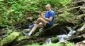 Man with mobile phone sits near small river in the forest HD Footage