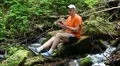 Man with mobile phone sits near small river in the forest HD Footage