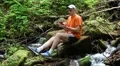 Man with mobile phone sits near small river in the forest HD Footage