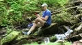 Man with mobile phone sits near small river in the forest HD Footage
