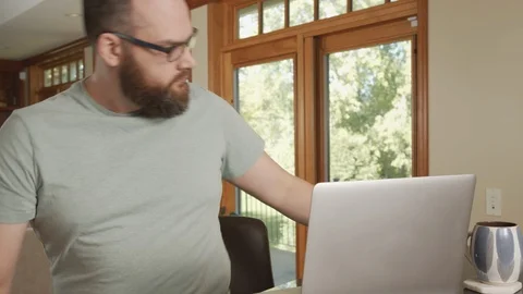 Man in modern home sits down at laptop computer with coffee and starts typing Stock Footage 125405061