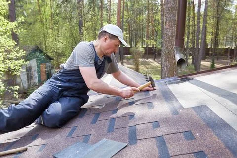 A man mounts soft bitumen shingles with nails and a hammer on the roof of a Stock Photos