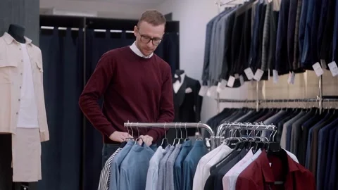 A man moves a hanger while choosing clothes in a clothing store.  Stock Footage 165958480