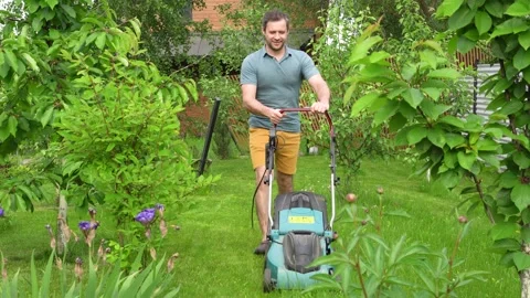 A man mows the grass with an electric lawn mower between young fruit trees. Stock Footage 155356065