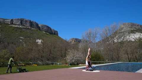 Man mows the grass. Girl doing yoga by the pool. Open air stretch. Quarantine Stock Footage 129608744