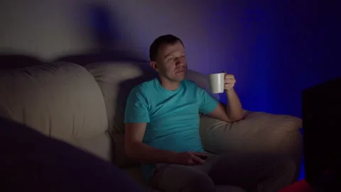 A man with a mug of hot tea sits at the TV late in the evening Stock Footage 131428673