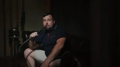 A man with a mustache and beard smokes a hookah in a hookah lounge. Video stock 253373554