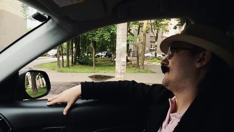 Man with mustache in dinner jacket, hat, glasses sitting in car and sing. Parody Video stock 75313750
