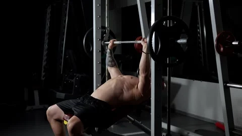 A man with a naked torso does bench press in a dark gym. Bodybuilder. Stock Footage 315860012