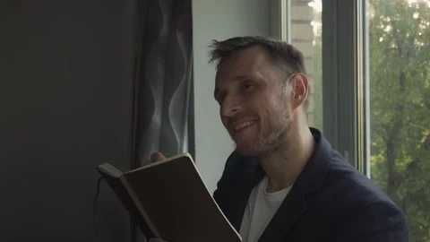 A man with a notebook in his hands composes poetry by the window Stock Footage 156142102