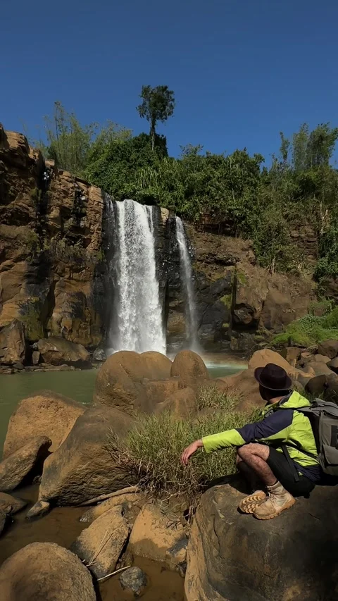 Man Observing Waterfall, Rocky River in Jungle, Sunny Vertical Wide Shot Stock Footage 324772526