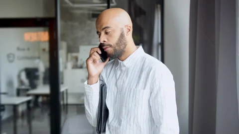 Man in office setting has phone conversation while standing near window, 스톡 동영상 335754546