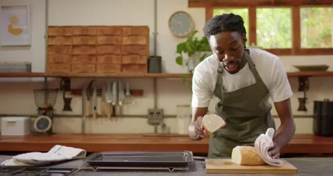 Man in olive-green apron lifting bread slice from loaf addressing camera while Stock Footage 342494837