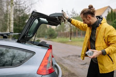 Man opens car trunk on a cloudy day while holding a water bottle near a dirt Stock-Fotos