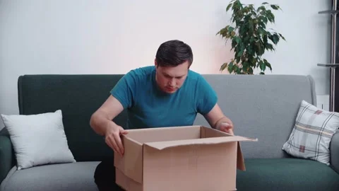A man opens a cardboard box while sitting on a home office couch. Stock Footage 145163219