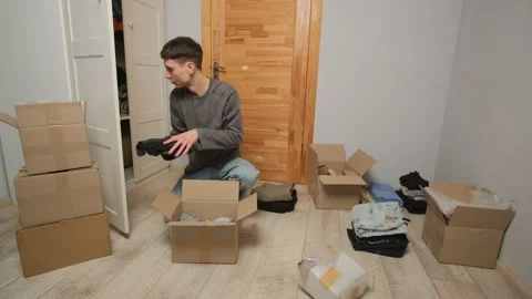 A man opens a large cardboard box, carefully taking out clothes and arranging Stock Footage 304636047