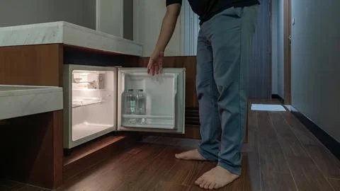 A man opens a small fridge built into a wooden cabinet in a dim hotel room,.. Stock Photos
