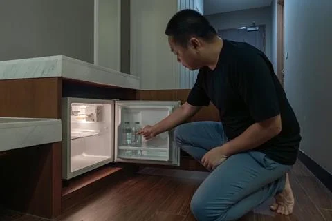 A man opens a small fridge built into a wooden cabinet in a dim hotel room,.. Stock Photos