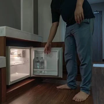 A man opens a small fridge built into a wooden cabinet in a dim hotel room,.. Foto stock