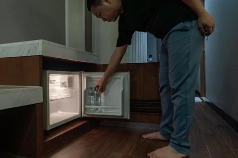 A man opens a small fridge built into a wooden cabinet in a dim hotel room,.. Stock Photos
