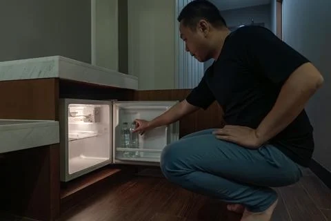 A man opens a small fridge built into a wooden cabinet in a dim hotel room,.. Stock Photos