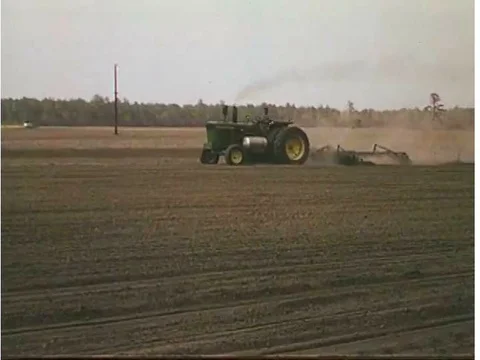A man operates a tractor pulling a seeding machine through a plowed field in the Stock Footage 74975811