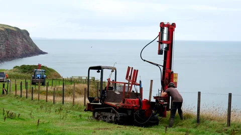Man operating tractor mounted fence post driver at seaside Stockbeeldmateriaal 249457338