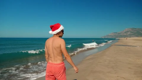 Man or guy in santa claus hat walking along sea on background of mountains. Stock Footage 253395669