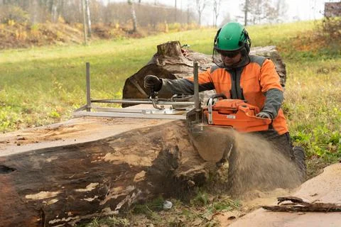 A man in an orange jacket on the side of a field cuts boards with an alaskan  Stock Photos