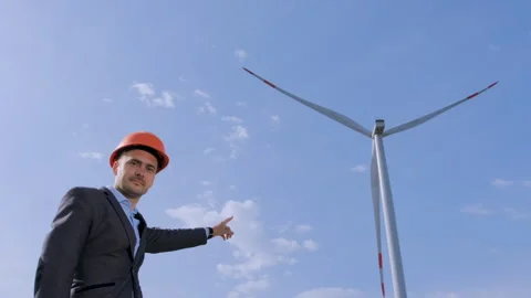A man in an orange protective helmet points his hand at the windmill. Stockbeeldmateriaal 134166955