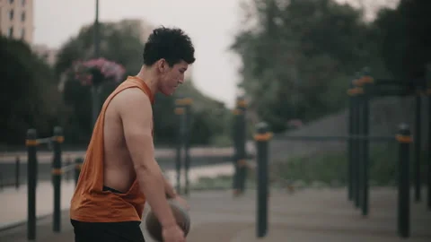Man in orange tank top playing basketball on outdoor court in urban park with Video stock 341875383