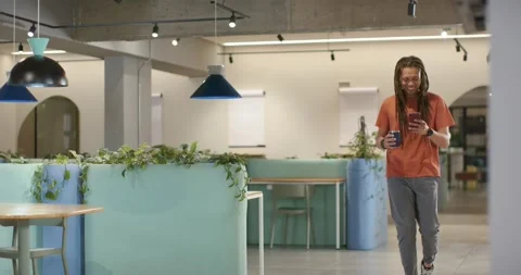 Man orange-tee coming from right into lounge walking checking work phone sipping Stock Footage 336305949