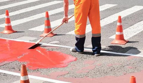 Man in orange work clothes repairs pedestrian crossing in morning, summer and Stock Photos