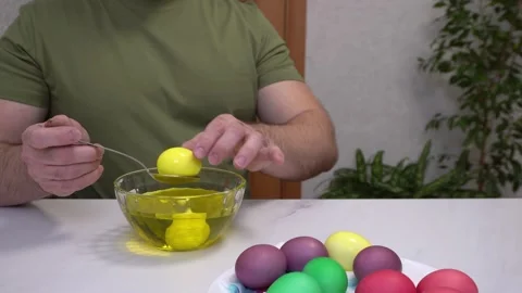 A man organizes colorful Easter eggs on a plate while sitting at home in the 動画素材 305025366