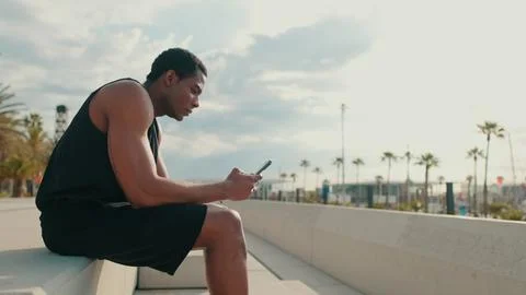 Man Outdoors Sitting On Bench Using Phone By Beach Boardwalk Stock Photos