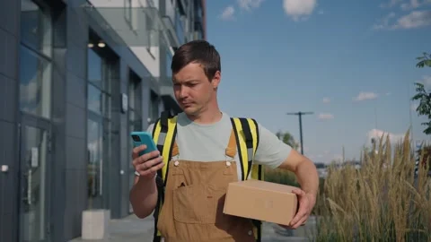 Man in overalls carrying a parcel uses a smartphone navigation app on a busy Video stock 317764536