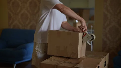 Man packing cardboard box using packing tape dispenser, preparing belongings for Stock Footage 294262023