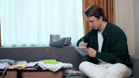 Man packing his suitcase for summer vacation travel, writing in a notebook to ch Stock Footage 342599291