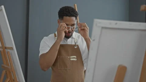 Man painter in studio wearing apron, holding paintbrush with hand on chin s.. Stock Footage 337439180