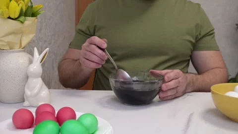A man paints Easter eggs at home, showing the process of lowering a white egg Stock Footage 305024635