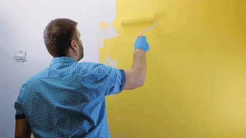 Man paints white wall with roller in his right hand in yellow. He is wearing Vidéo 123098920