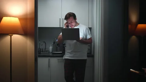 Man in pajamas stand in kitchen talking on smartphone and working on laptop Stock Footage 194568766