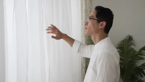 Man parting curtains and looking out of window Stock Footage 251839956