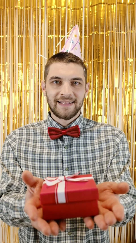Man in a party hat and red bow tie smiles and offers a red gift box with a white Stock Footage 342621933