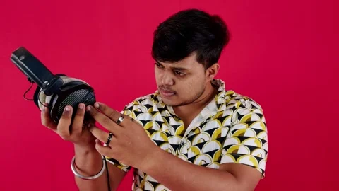 Man in patterned shirt examining headphones against a red background Stock Footage 262361704
