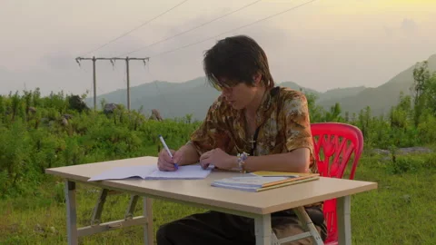 Man In A Patterned Shirt Sitting At A Desk Outdoors, Writing In A Notebook Stock Footage 342866255