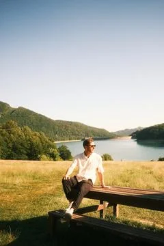 A man is peacefully sitting on a bench while overlooking a serene lake Stock Photos