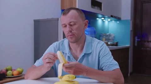 A man peels and eats a banana sitting at a table, kitchen background 스톡 동영상 331137612