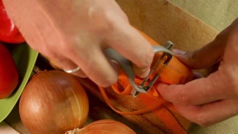 Man peels carrot using metal peeler on wooden cutting board with onions and Stock Footage 318781953
