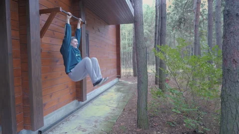 Man performing hanging knee raises outdoors near wooden house Stock Footage 334331535